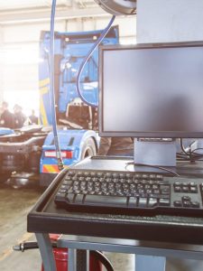 Monitor and keyboard at a stationary service point of trucks in a large garage and people in the background. Professional computer diagnostics of technical condition and malfunctions. Checking engine